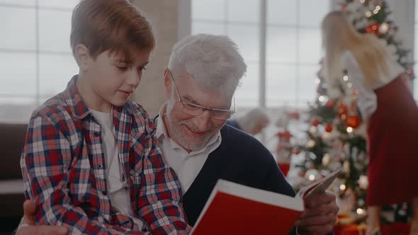 Grandad Reading a Book with Grandson Sitting on a Chair at Christmas Celebration alt