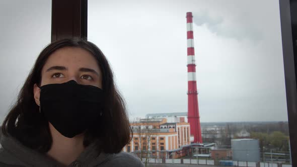 Teen Girl in a Protective Mask on the Background of Smoke From a Pipe Through the Glass