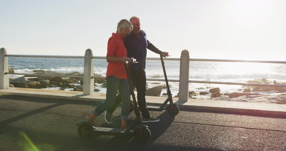 Senior couple walking next to electronic scooter alongside beach alt