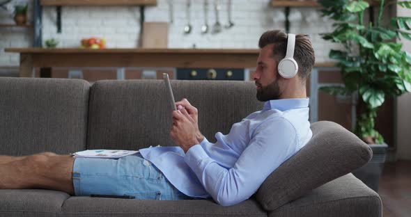 Handsome Businessman in Headphones Lying in Cozy Couch Looking Document Paper in Modern Kitchen alt