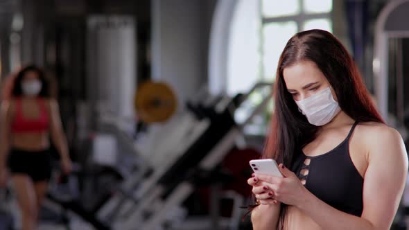 А Girl in the Gym in a Face Protective Mask Uses a Smartphone. In the Background, Other Athletes