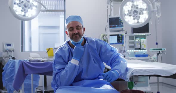 Portrait of caucasian male surgeon wearing lowered face mask sitting in operating theatre smiling alt