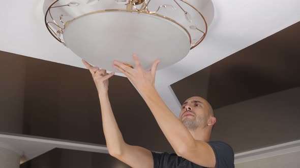 A Man Standing on a Stepladder Tries on a Ceiling Lamp on a Chandelier alt