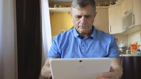 Portrait Of A Senior Man At The Blue Shirt Sits And Types A Tablet Pc At Home alt