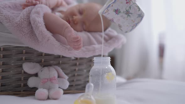 Pouring Milk in Baby Bottle with Blurred Infant Sleeping at Background in Baby Basket alt