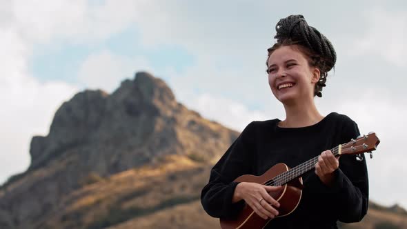 Young Laughing Woman with Dreadlocks Tied Up to Her Head Playing Ukulele in Mountains alt