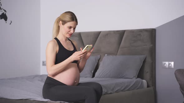 Pregnant Woman Sits on the Bed in the Bedroom and Holds a Smartphone in Her Hands She Smiles at the