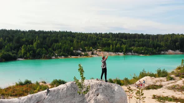 Landscape of a Light Blue Lake Surrounded By Forest - a Successful Man Standing on a Rock and Raises alt