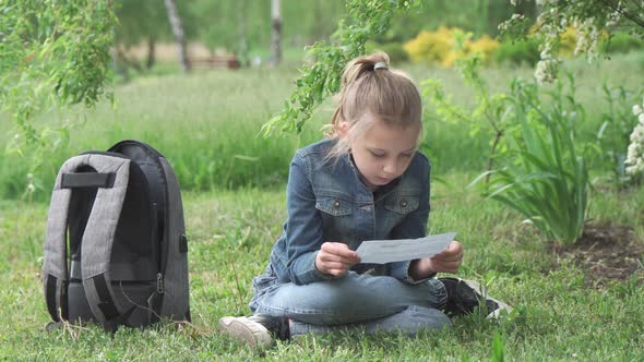 A girl of school age sits in a park on the grass and reads carefully.