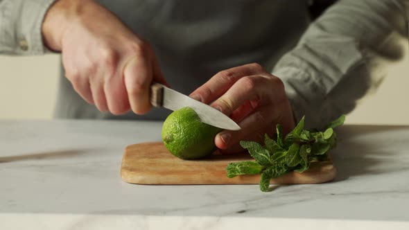 Bartender is Cutting a Juicy Green Lime on a Chopping Board with a Knife and a Sprig of Mint is alt