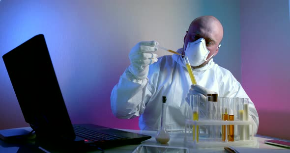 Man in a White Coat, Respirator and Gloves Is Sitting at a Table with a Gadgets. He Holds a Tube alt