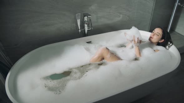 Cute Girl Lying in Bath Indoors. Sweet Lady Enjoying Bubble Bathtub. alt