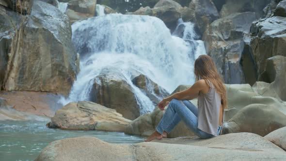 Barefoot Girl Sits and Looks at Waterfall on Rocks