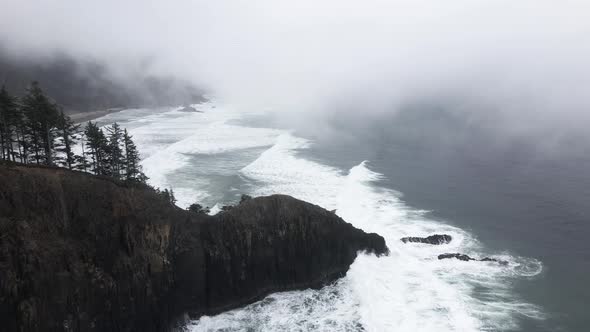 Descending on a fog shrouded tree lined ridge plunging into a jagged Oregon coastline, aerial alt