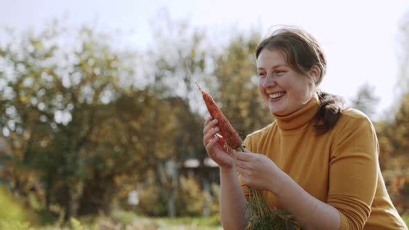 Happy Simple Woman Sits in Garden and Examines Fresh Raw Carrots alt