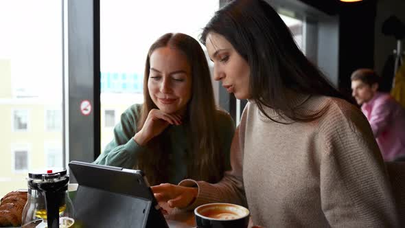 Two Young Beautiful Girls in a Cafe are Looking at a Tablet