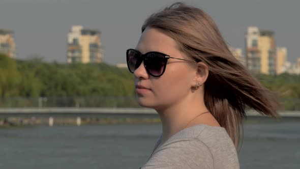 Portrait of a Beautiful Blonde Girl in Sunglasses with Waving Hair Looking at the River in the Park alt