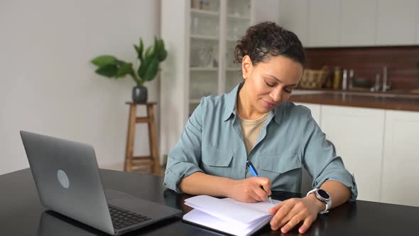 Smiling Female Using Laptop for Watching Webinars Sits at Desk in Home Office and Taking Notes