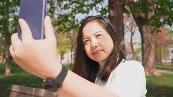 Close up Asian woman sitting on a chair at a park and using her smartphone to take a selfies alt