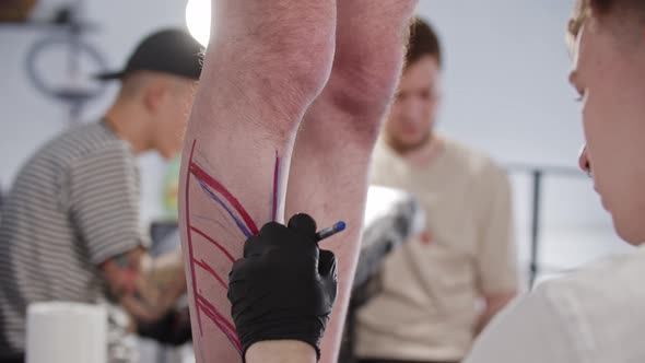 Young Man Tattoo Artist Drawing a Sketch on the Leg on His Client with a Blue Pen alt