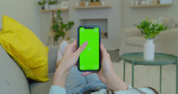 Young Woman Home Lying on a Couch with Green Screen Smartphone. Girl Using Touchscreen Mobile Phone alt