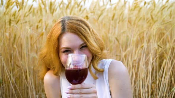 Happy red haired woman drinking wine at picnic in wheat field alt
