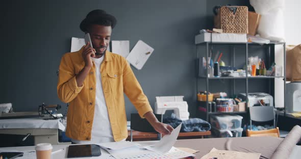 Trendy Tailor Talking on Mobile Phone and Looking at Papers Working in Studio alt