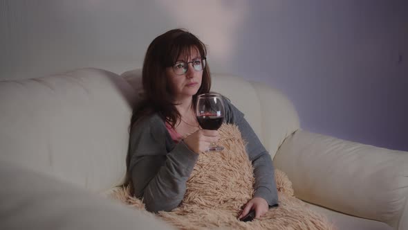 Sad Brunette Woman Relax at Home with a Glass of Wine on the Sofa and Watches Tv in the Evening