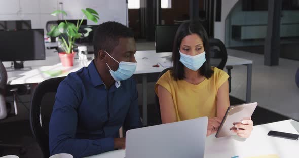 Diverse male and female business colleagues wearing face masks sitting at desk using tablet alt