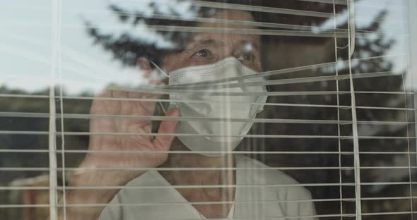 Elderly Woman Wearing Face Mask Looking Through Window Blinds