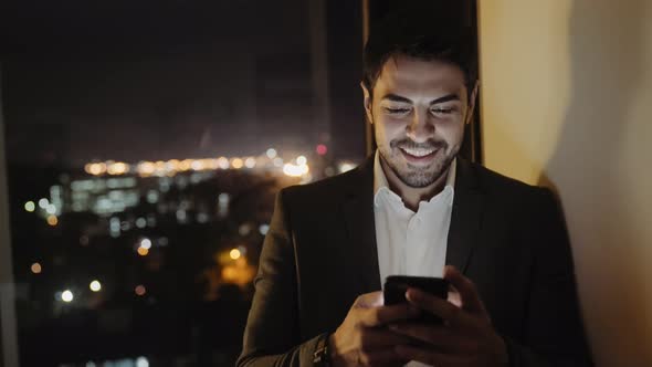 A Handsome Young Man in a Suit Smiles While Texting Using his Smartphone at Night By Window