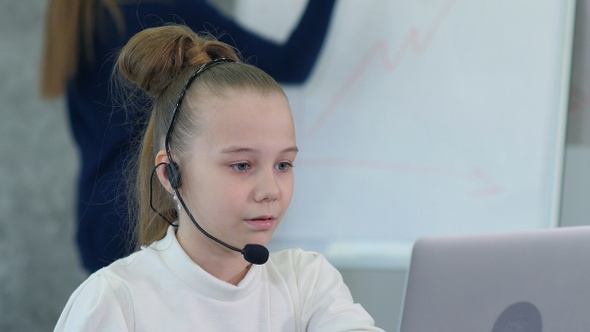 Cute little girl in headset sitting at table with laptop