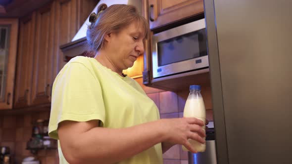 Middle-aged woman opens refrigerator at night and drinks milk from a plastic bottle. alt