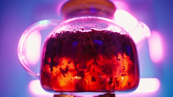 Transparent Teapot with Tea Leaves Boils on Gas Burner Against Neon Backlight alt