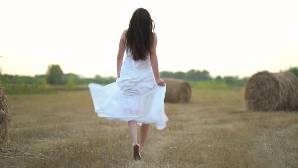 Beautiful Pregnant Woman Running in Wheat Field with Haystacks at Summer Day