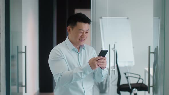 Positive Middle Aged Asian Businessman Typing on Smartphone Chatting with Colleagues and Smiling at alt