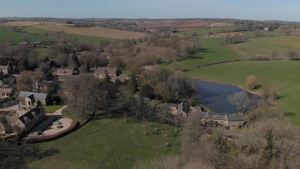 Aerial View Of Quaint English Cotswold Village And Lake Disappearing Behind Trees 4K Cine D