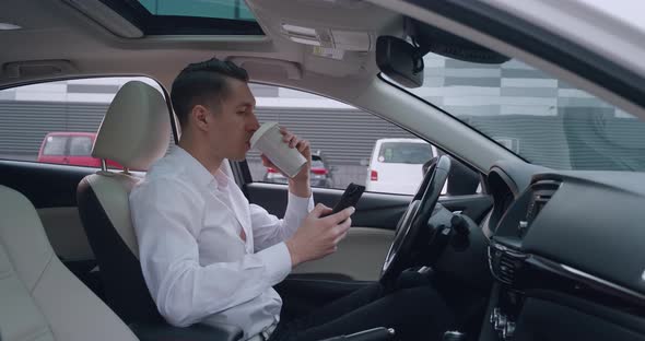 Portrait of Young Man Entrepreneur Sits in the Driver's Seat in a Car and Uses Smartphone for alt