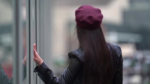 a Brunette with Long Hair in a Black Leather Jacket and a Burgundy Cap Walks Away From the Glass alt