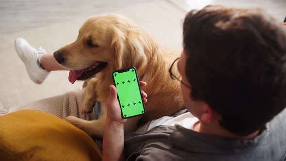 Brunette Man Wearing Glasses Sitting on Sofa with Golden Retriever Using Smartphone for Buying alt