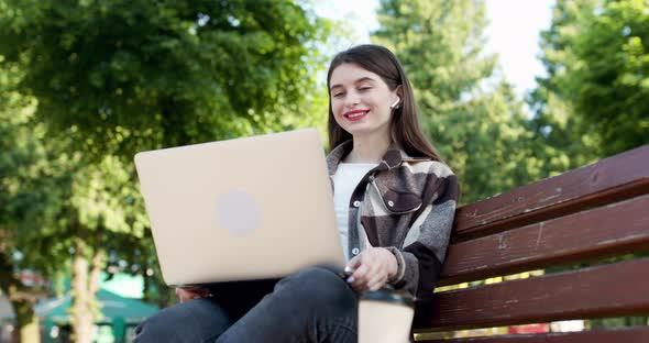 Girl is Waving Her Hand to Webcam on Laptop While Sitting on Park Bench alt