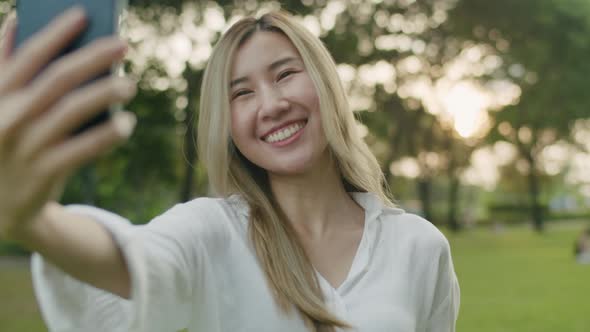 Smiling Asian female taking selfie photo on smartphone at a public park during sunset.