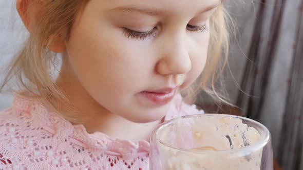 A Cute Preschooler Girl in a Pink Blouse Drinks Milk Cocoa From a Glass Holding It with Both Hands