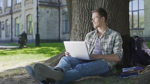 Senior Student Working on His Project on Laptop Sitting Under Wide Tree, Campus alt