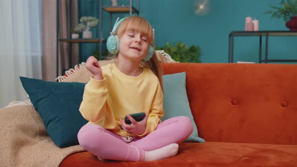 Child Girl Kid with Smartphone in Headphones Dancing While Listening Music at Home Alone on Sofa alt