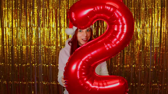 Woman in Santa Hat Holds Red Figure Two on Background of Shimmering Golden Tinsel alt