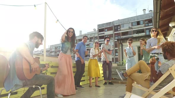 Musician playing guitar at rooftop party, people dancing and taking photos alt
