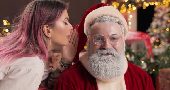 A Young Woman Whispers Something in the Ear of a Surprised Santa Claus Sitting in a New Year's