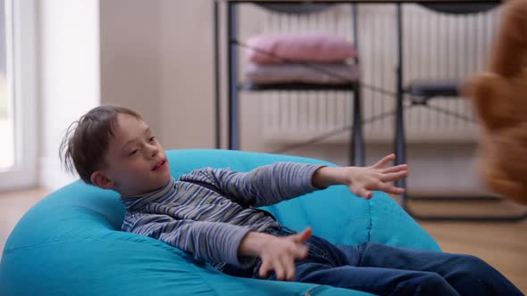 Joyful Autistic Boy Lying on Comfortable Bag Chair at Home Throwing Catching Teddy Bear alt