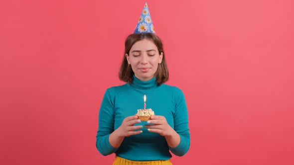 Young Woman in Birthday Hat Blowing Out Candle on Cake, Isolated on Red Background. alt